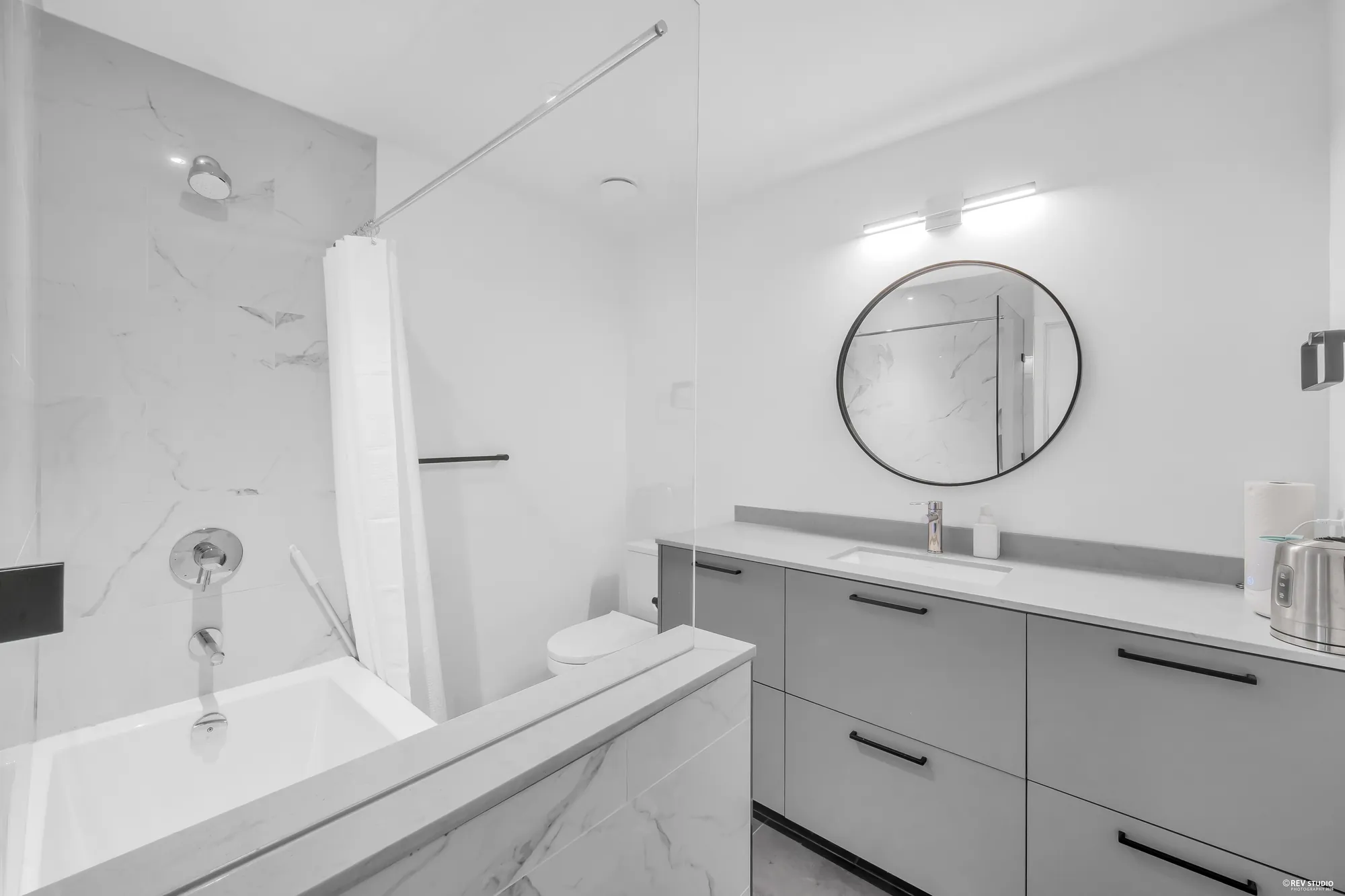 The upper bathroom: a tub with a shower and a curtain, marble-look tile, a grey vanity with a round mirror