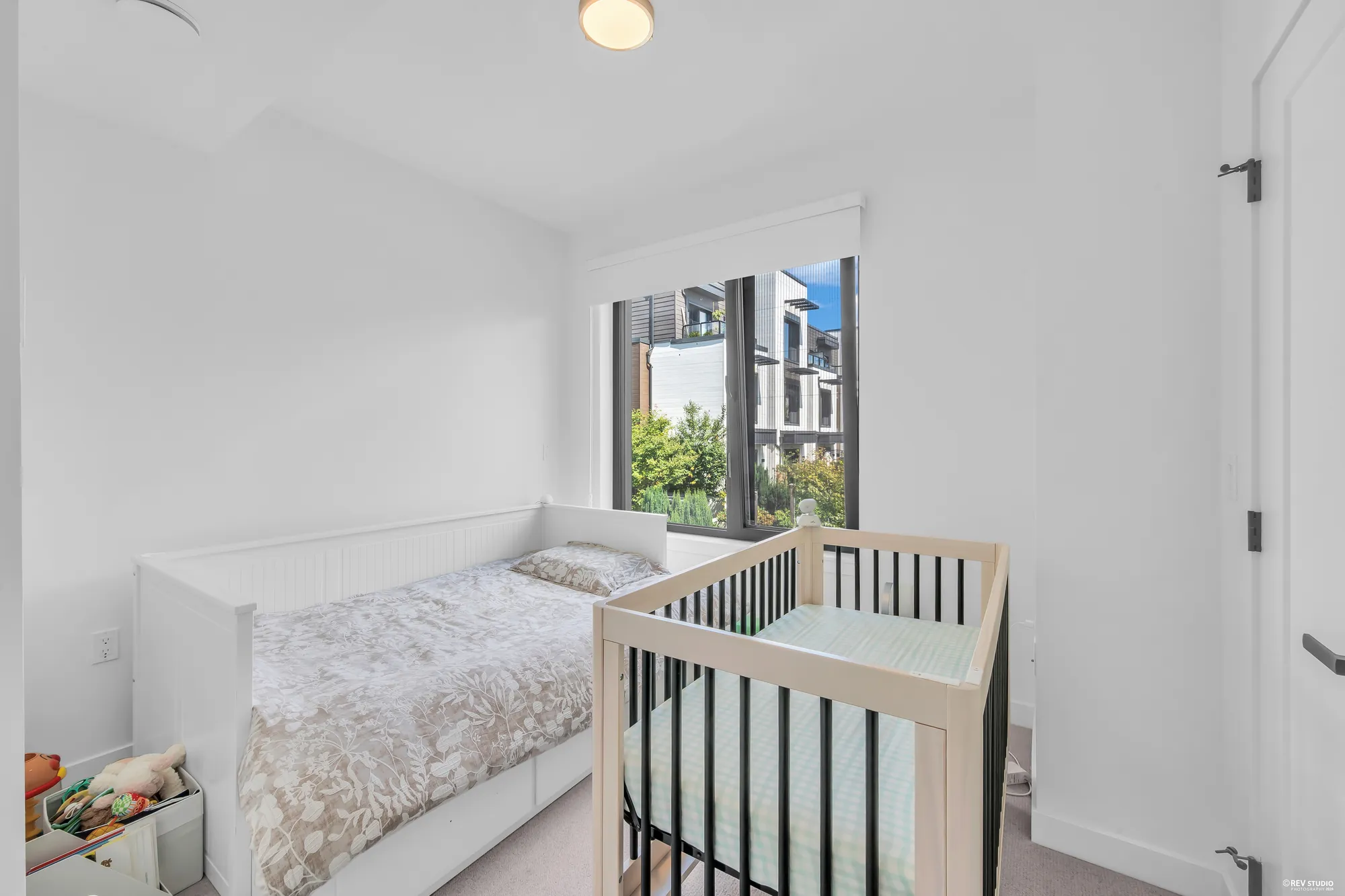 The second upper bedroom: a white daybed and a crib, a wide window onto a tree and the townhomes across the way