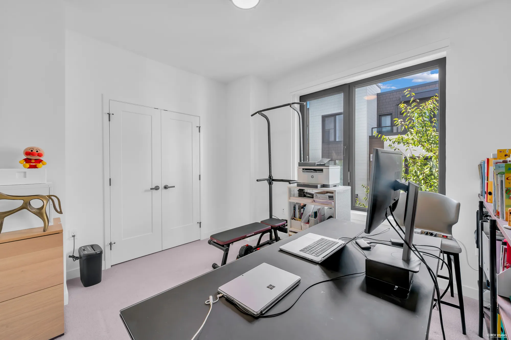 The larger upper bedroom, set up as an office: a double closet, a wide window onto a tree and the townhomes behind, a desk with a monitor and laptops, a weight bench and a pull-up stand