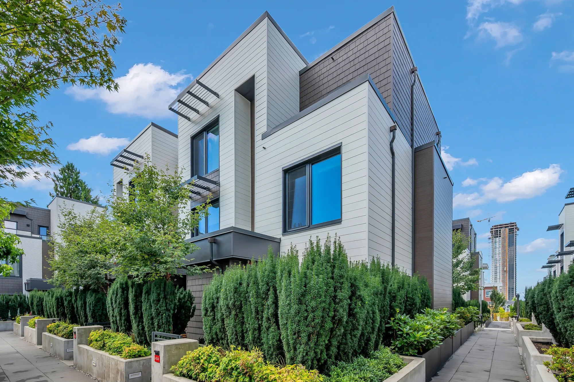 The corner of the building at 404-606 Foster Avenue: white siding and a dark shingled upper storey, a tall window over clipped yew hedges in a concrete planter, paved walkways on both sides