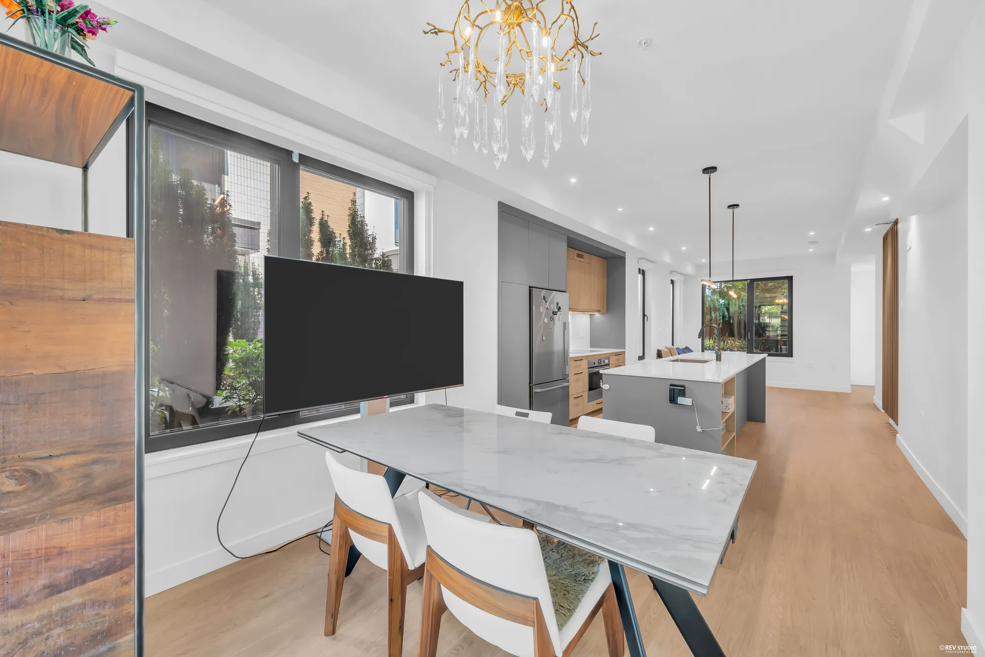 The dining room toward the kitchen: the table and chairs, the chandelier, a window onto the hedge, the island and the living room beyond