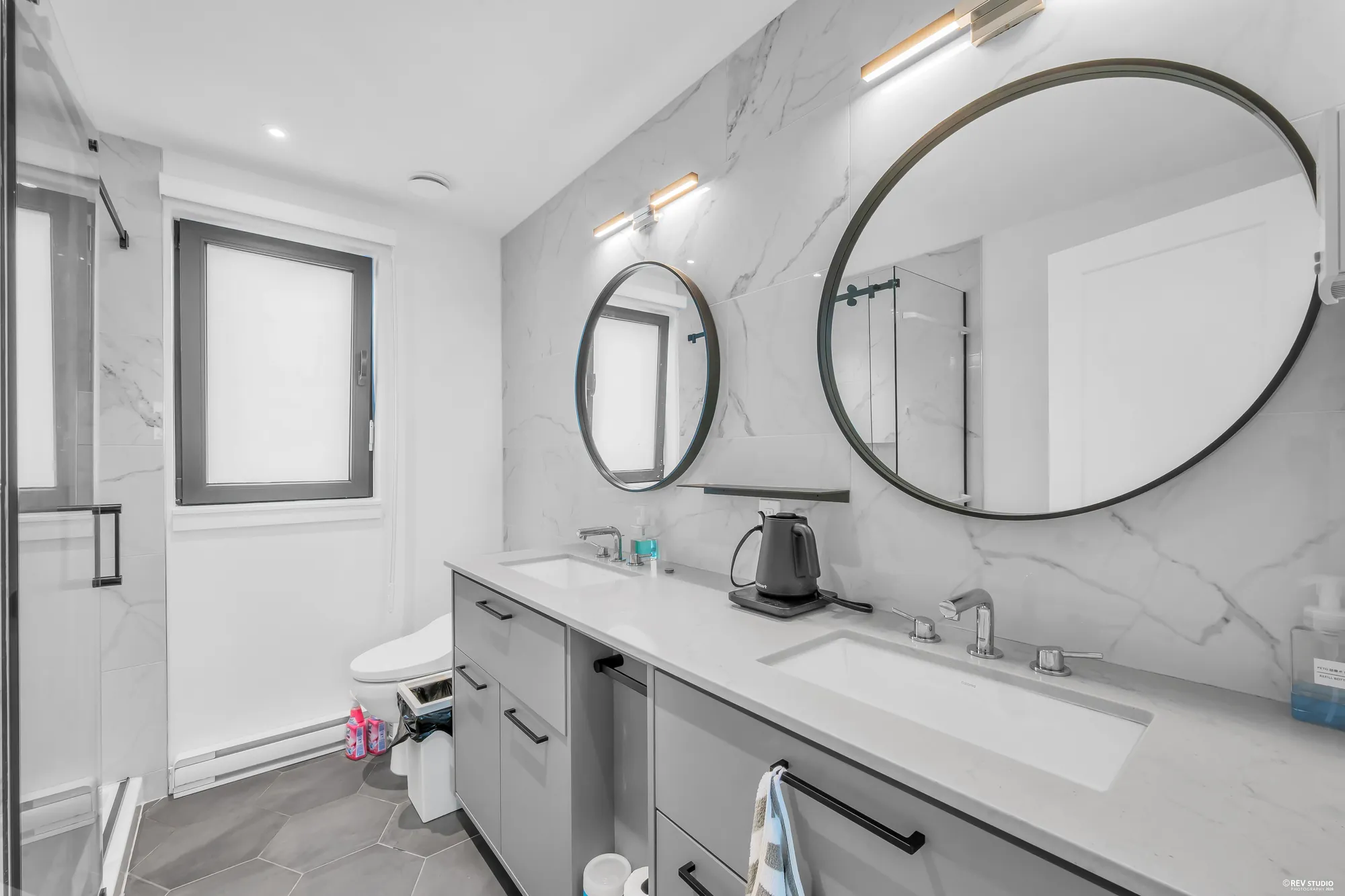 The primary bathroom: a long grey vanity with two sinks and two round mirrors, marble-look tile, a frosted window, a toilet
