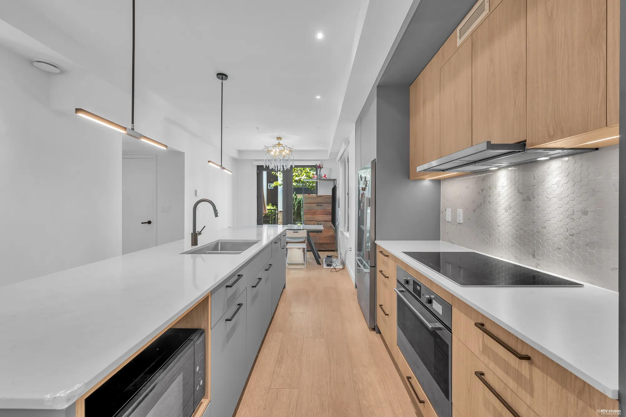 The kitchen: wood-grain upper cabinets, a hexagon-tile backsplash, a black glass cooktop under a pull-out hood, a wall oven below it, the grey island with the sink