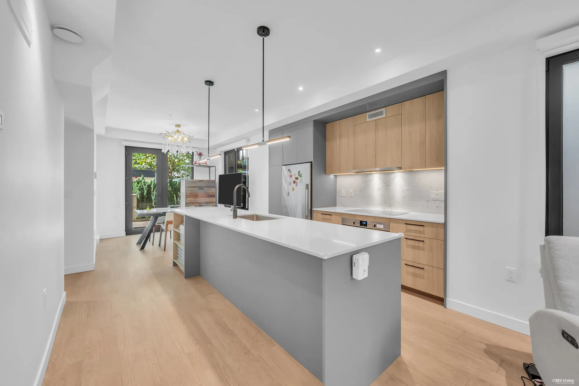 The kitchen from the living room: the grey island, the wood-grain cabinets and the cooktop, a stainless fridge, the dining room beyond