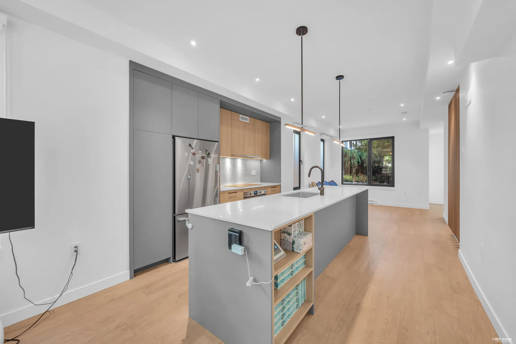 The kitchen from the dining room: the island with open shelves at its end, a stainless fridge in a wall of tall grey cabinets, the living room beyond