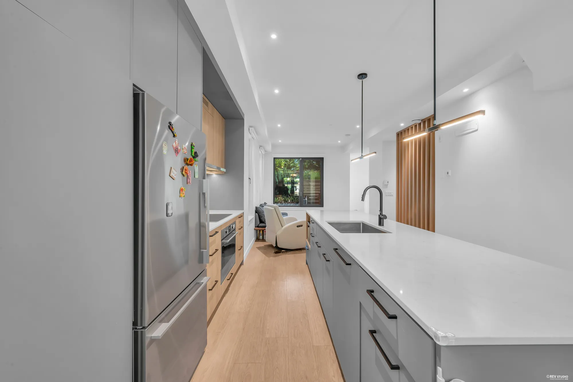 The stainless fridge in the wall of tall grey cabinets, the island at the right, the living room and its window beyond