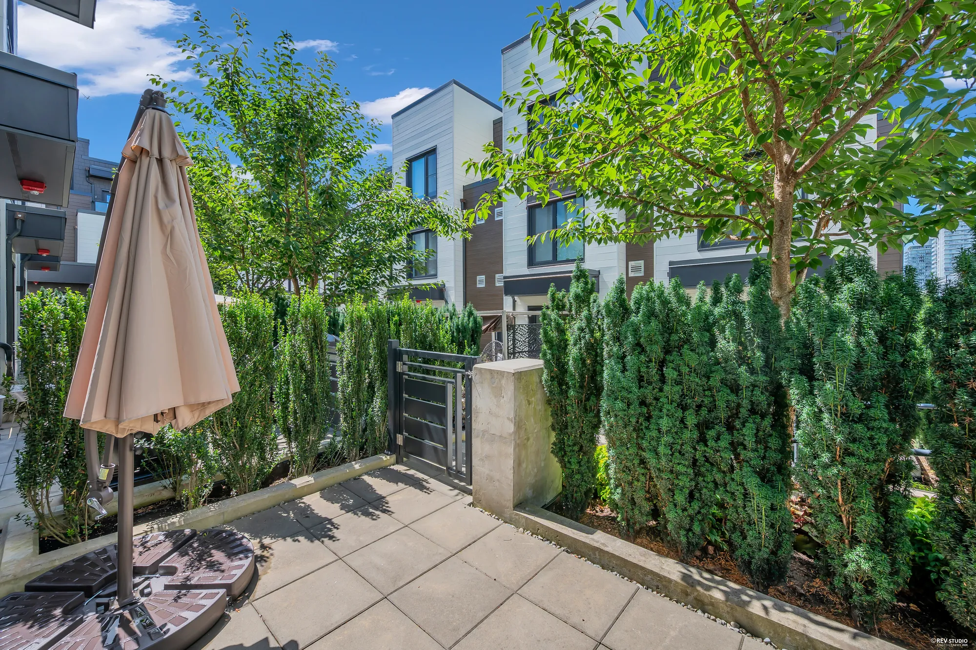 The back patio: pavers, a folded market umbrella on a base, a steel gate to the walkway, yew hedges and a tree, townhomes across the way