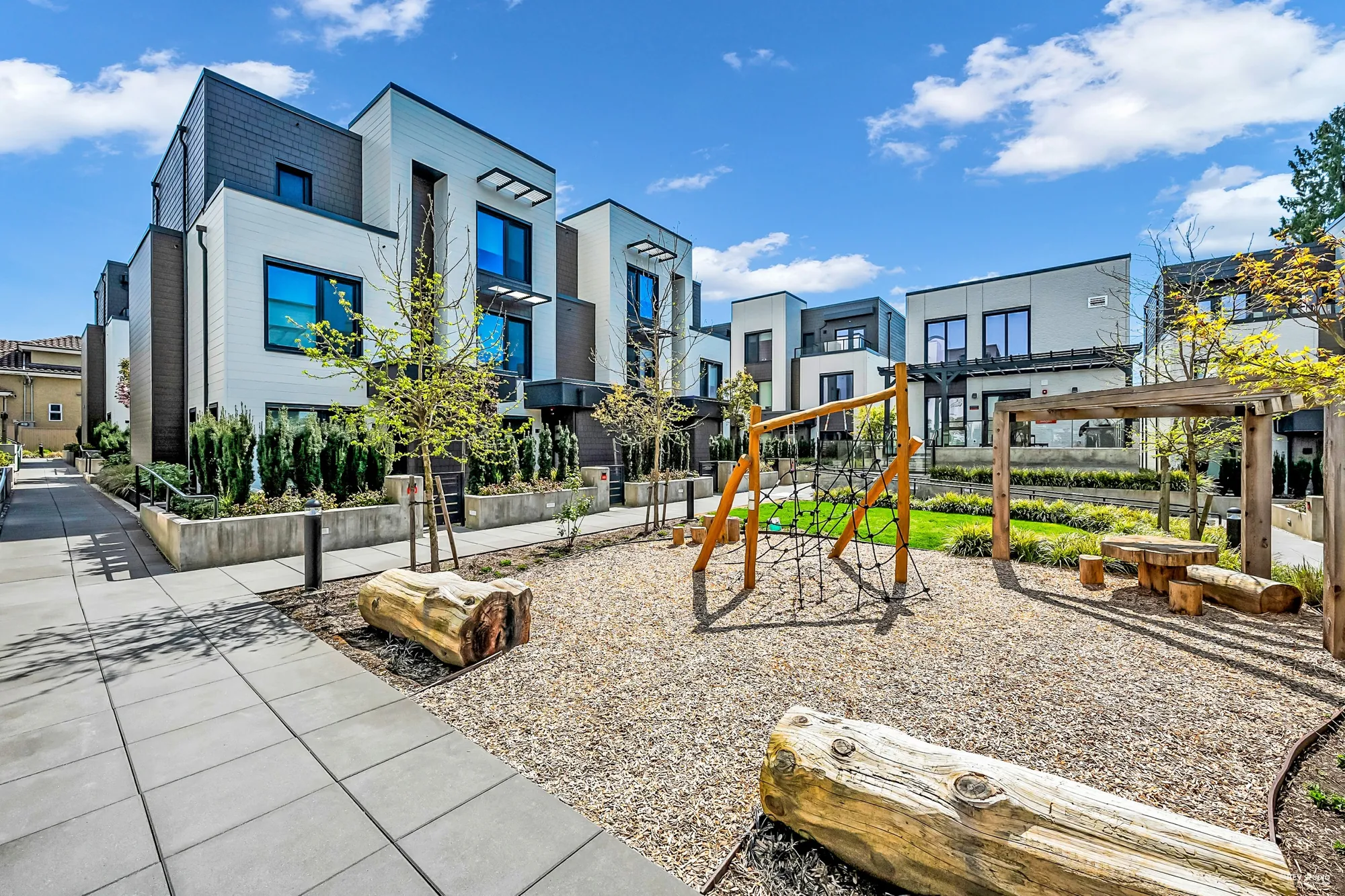 The play area inside the complex: a rope climbing frame, a timber pergola, log seats on wood chips, a strip of lawn, townhomes around it