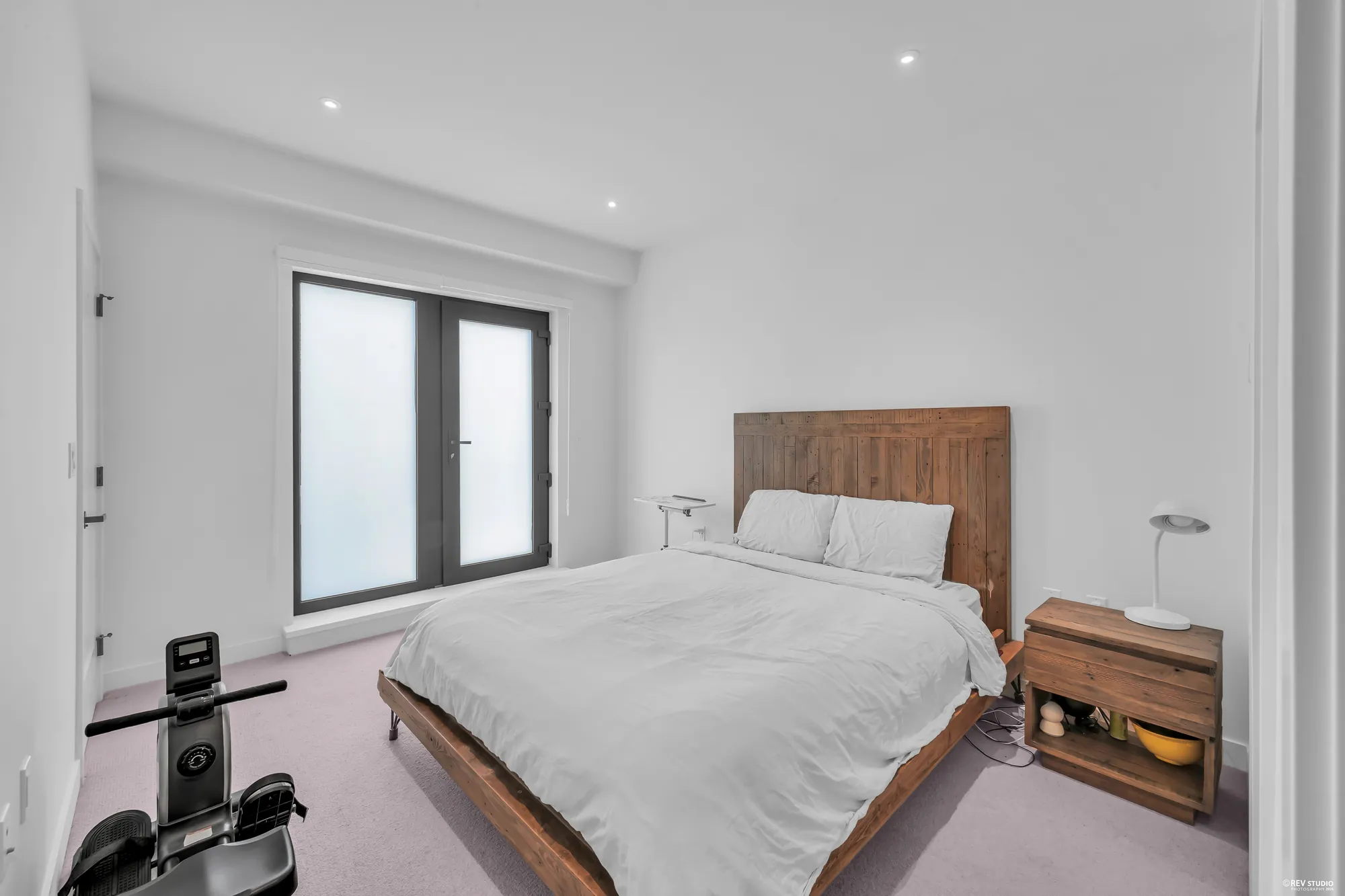 The primary bedroom toward the balcony: a glass door and a fixed glass panel in the far wall, the bed and a nightstand, a rowing machine
